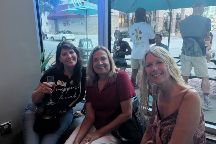 Three women smiling indoors at a cafe with a window view of people outside.