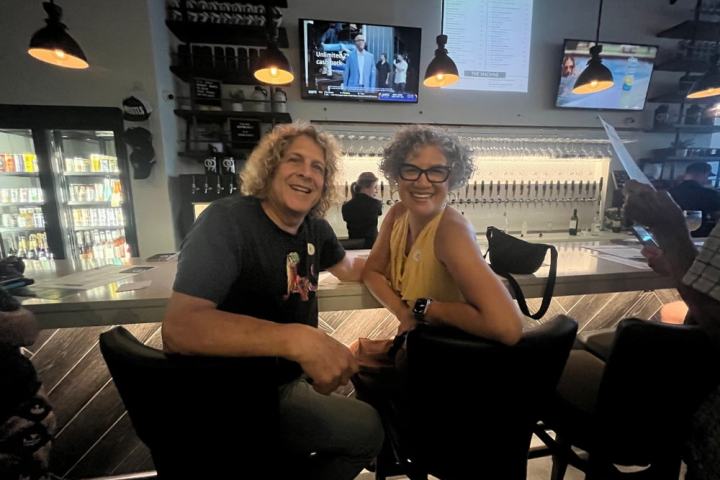 Two people smiling at a bar with hanging lights and TVs in the background.