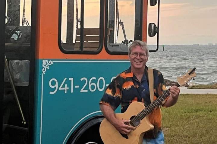 Man with guitar standing near colorful bus by the waterfront at sunset.