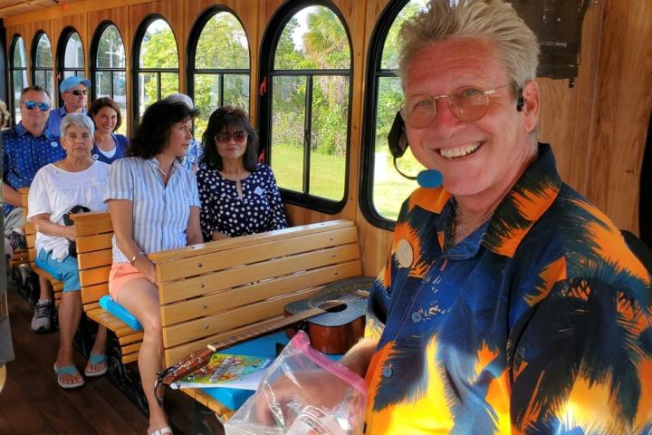 Smiling tour guide in a colorful shirt on a trolley with seated passengers enjoying the ride.