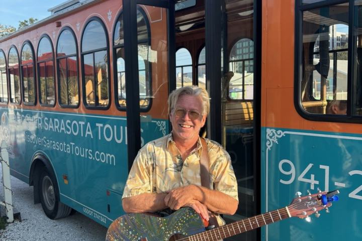 Smiling person with guitar stands in front of an orange tour bus.