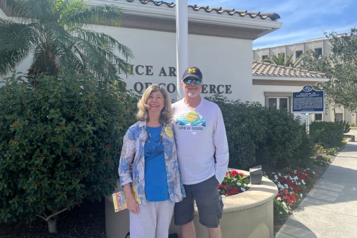 Two people standing outside a building with a palm tree and flowers.