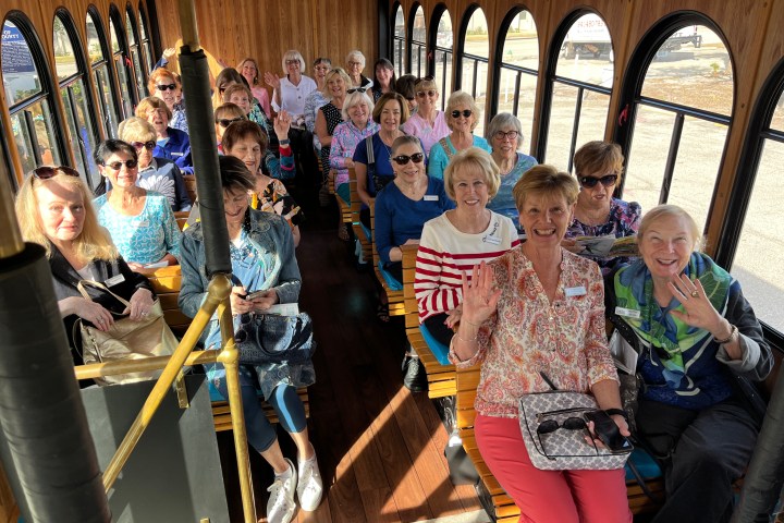A group of smiling people seated inside a wooden trolley, waving at the camera.