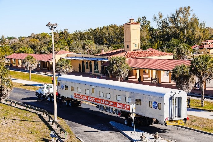 Circus train car parked near a building with palm trees in a sunny setting.
