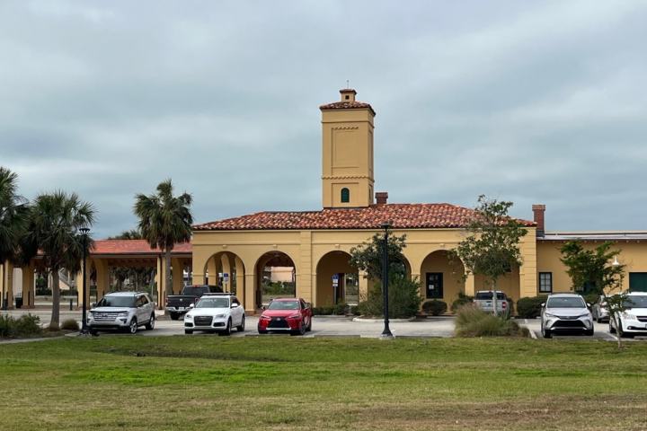 Spanish-style building with tower, cars parked in front, and palm trees around.