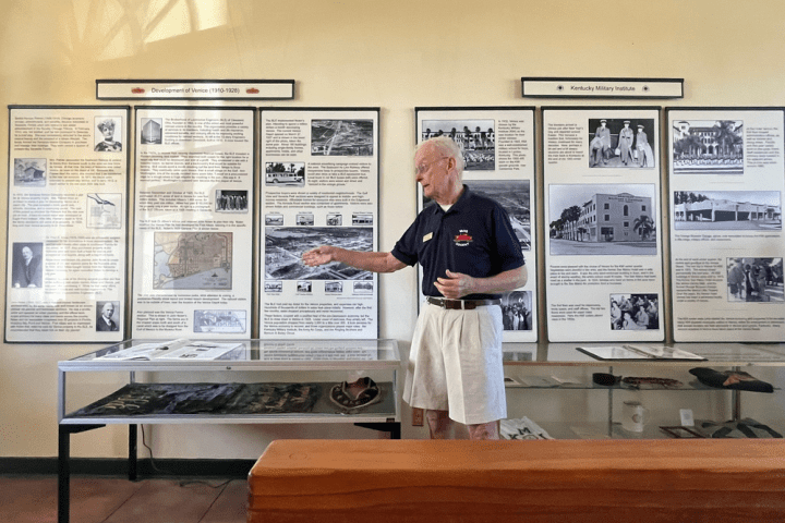 Man in a museum gestures in front of historical display panels and a glass case.