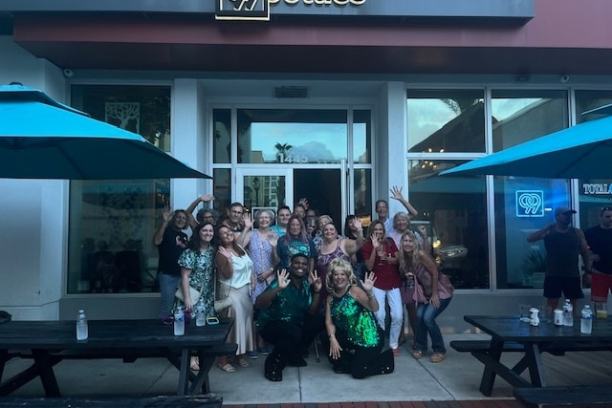 Group of people posing and smiling outside a store with a red awning and glass doors.