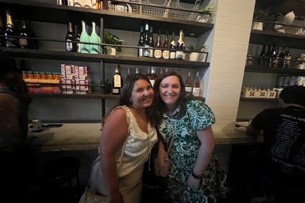 Two women posing in front of shelves with bottles and plants in a bar.