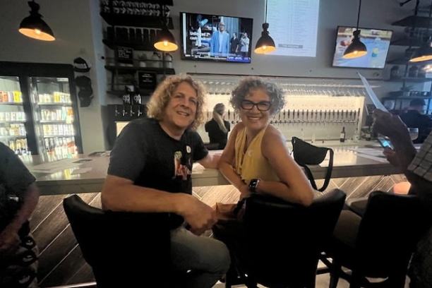 Two people sitting on barstools in a dimly lit bar with TV screens overhead.