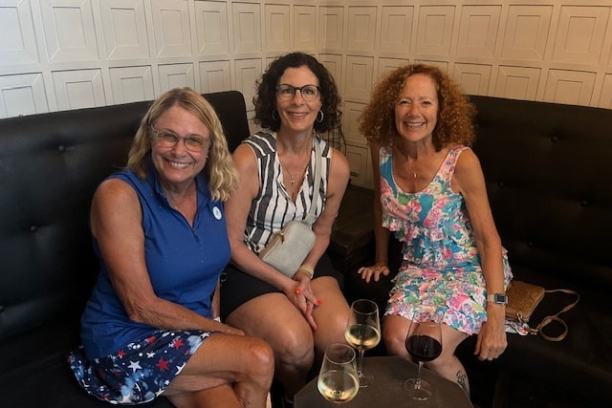 Three women sitting on a sofa in a room with wine glasses on the table.