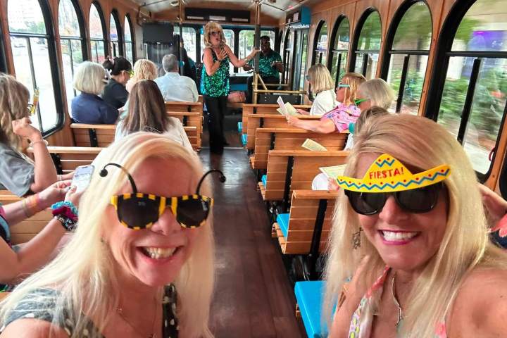 Two women wearing festive sunglasses pose on a trolley with other passengers seated.