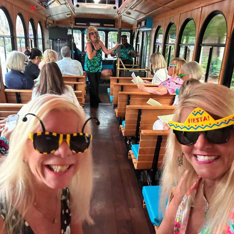 Two women wearing festive sunglasses pose on a trolley with other passengers seated.