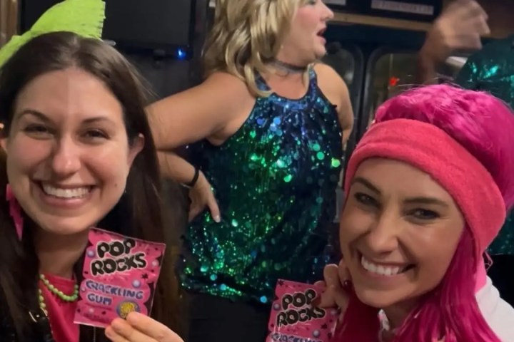 Three women smiling, one holding popping candy, wearing colorful outfits in a party setting.