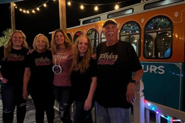 Five people posing in front of a decorated trolley at night, with string lights overhead.