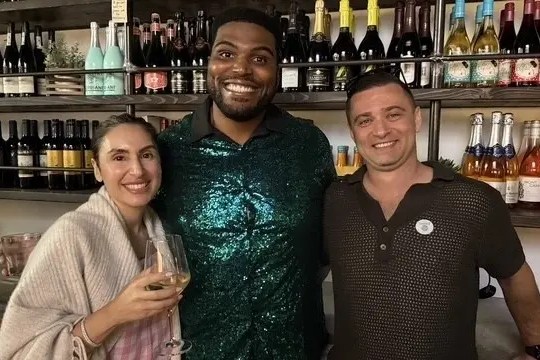 Three people smiling in a wine bar with shelves of wine bottles behind them.