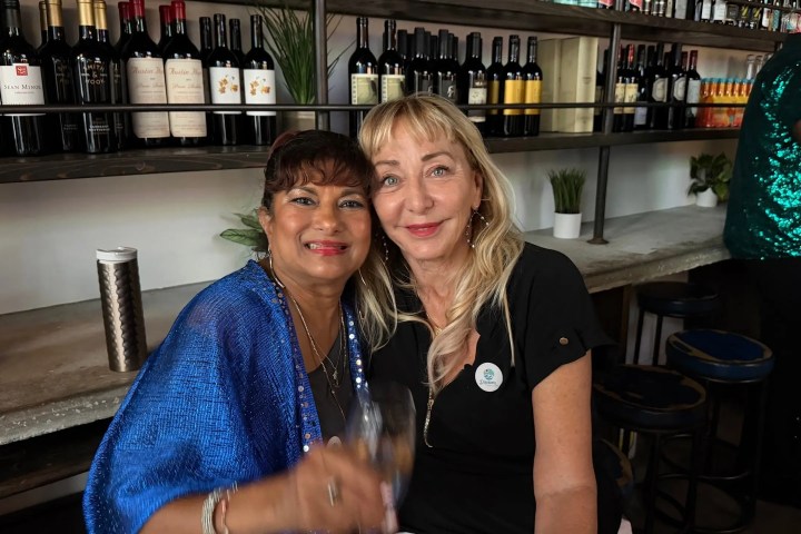 Two women smiling at a bar with wine bottles on shelves behind them.