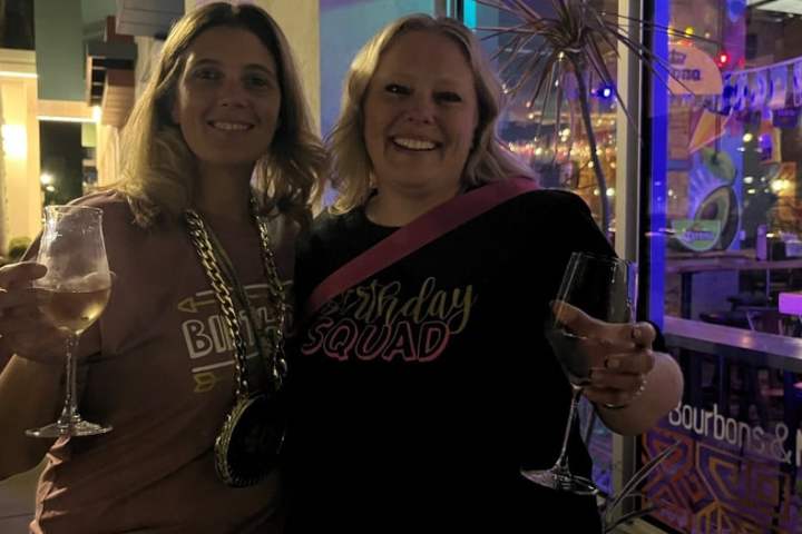Two women smiling at night, holding wine glasses outside a decorated venue.