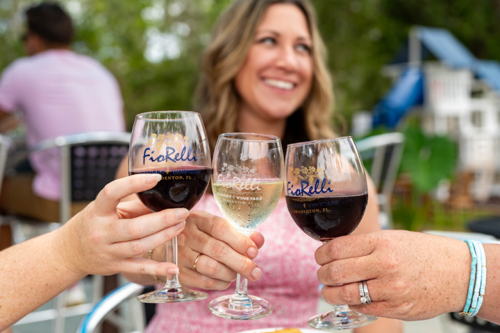 Three people clink wine glasses outdoors, smiling in background.