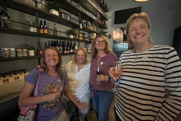 Four women smiling with drinks, standing in a modern brewery or bar with shelves of bottles in the background.