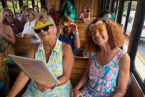 Two women in colorful dresses wearing novelty sunglasses on a bus with others.