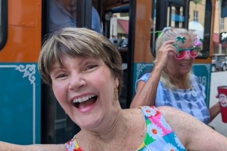 Smiling woman in floral dress near colorful trolley, another woman in novelty glasses in background.
