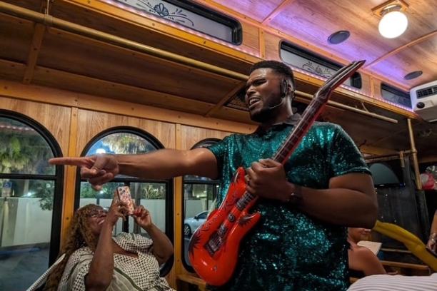Man in a shiny green shirt playing a red guitar and singing, with a seated audience inside a wooden trolley.