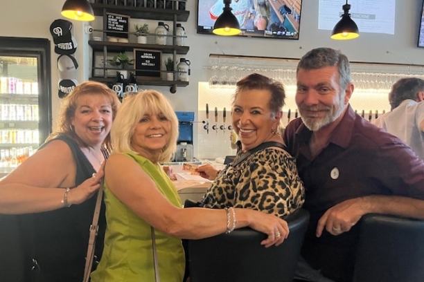 Four people smiling and posing together at a bar with TV screens above.