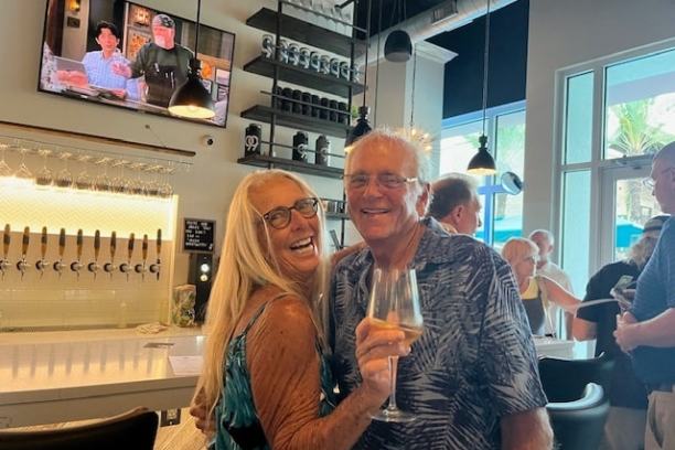 Smiling couple with wine glasses at a bar with TV and taps in the background.