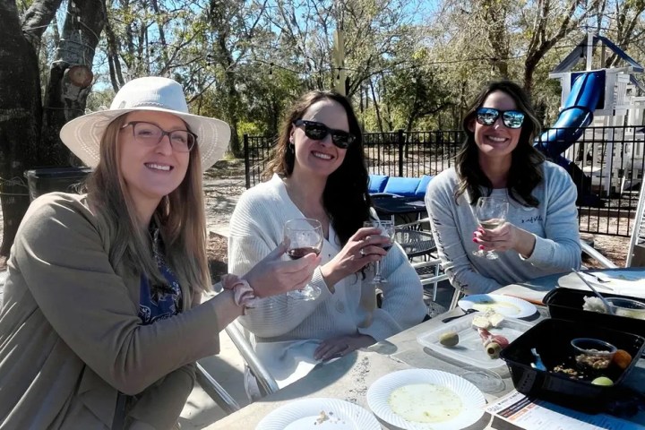Three women outdoors enjoying drinks and food at a picnic table.
