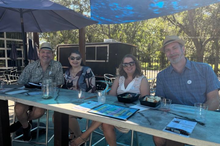 Four people sitting at a restaurant table outdoors, under umbrellas, smiling with drinks and food.