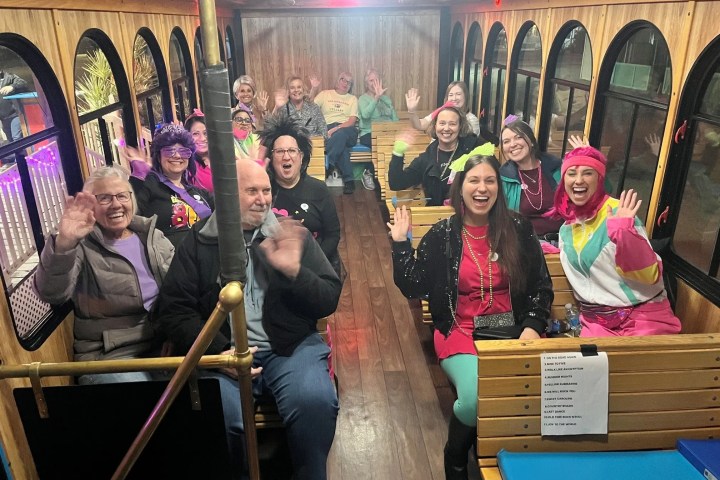 Group of people in colorful outfits smiling and waving on a trolley.