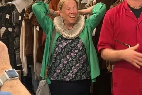 Woman in costume closet laughs with hands on head, man in red gestures beside her.
