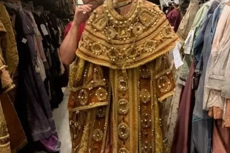 Person holding ornate, gold-embroidered robe in a costume storage room.
