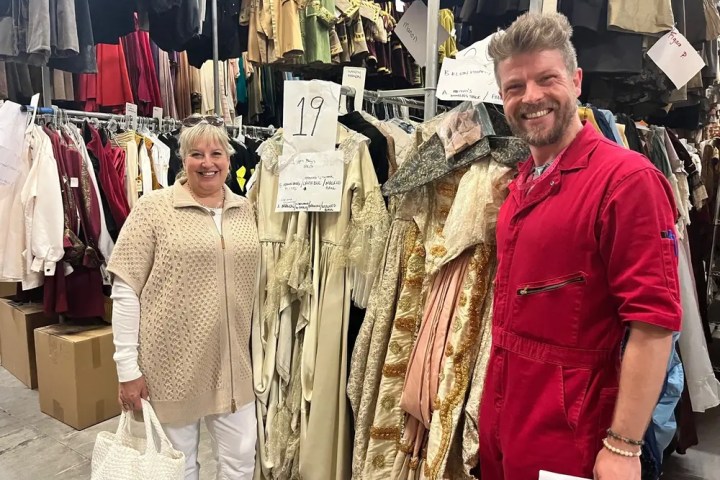 Two people in a costume storage room, surrounded by hanging clothes and outfits.