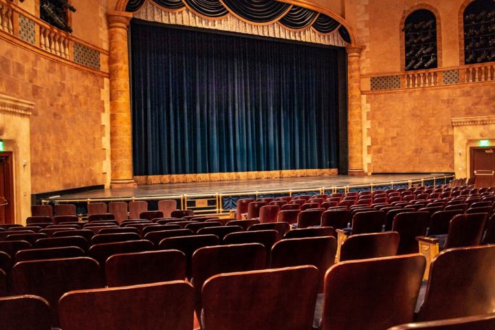 Empty theater with red seats, blue curtain stage, and ornate interior design.
