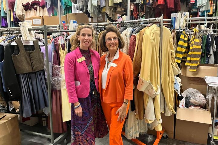 Two women in colorful outfits stand in a costume storage room.
