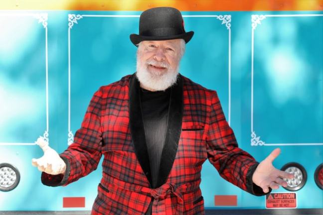 Smiling man in plaid jacket and bowler hat gestures in front of a colorful background.