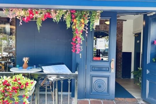 Blue cafe entrance with hanging flowers, menu stand, and outdoor table with chairs.