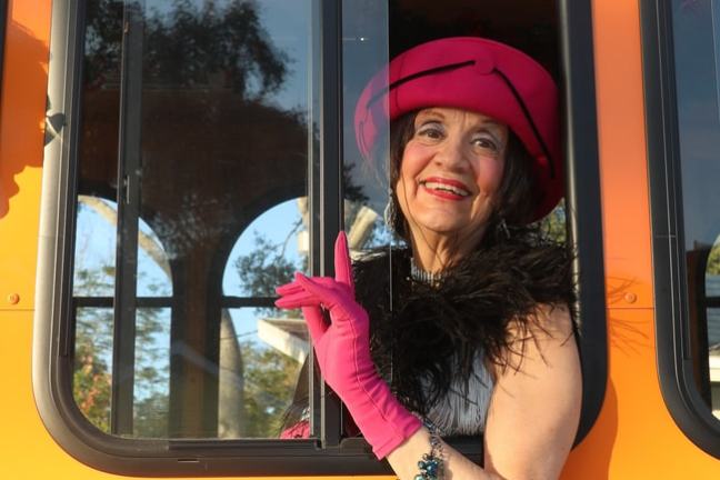 Woman in a bright pink hat and gloves smiles from a bus window.