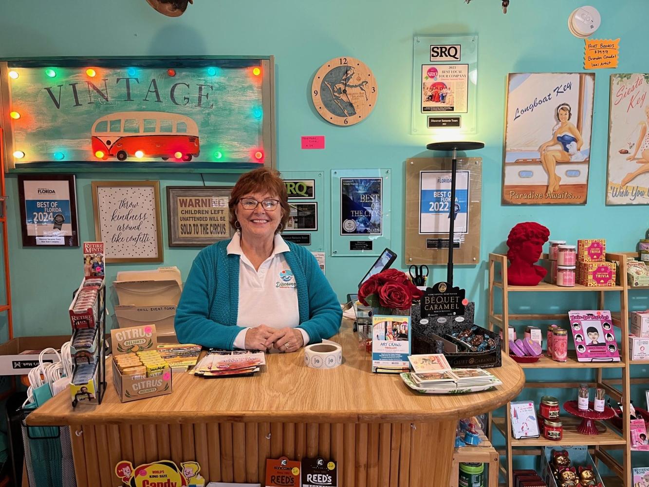 Smiling woman behind a retro-themed shop counter, surrounded by vintage decor and products.