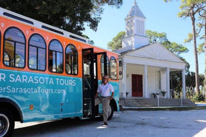 Person exiting tour bus near white church with trees in background.