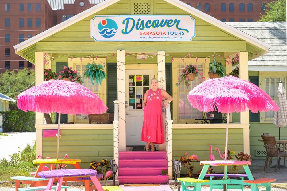 Woman in pink dress stands at doorway of colorful tour office with pink umbrellas and chairs.