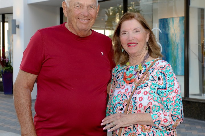 Smiling couple standing outside a building, man in red shirt, woman in patterned dress.