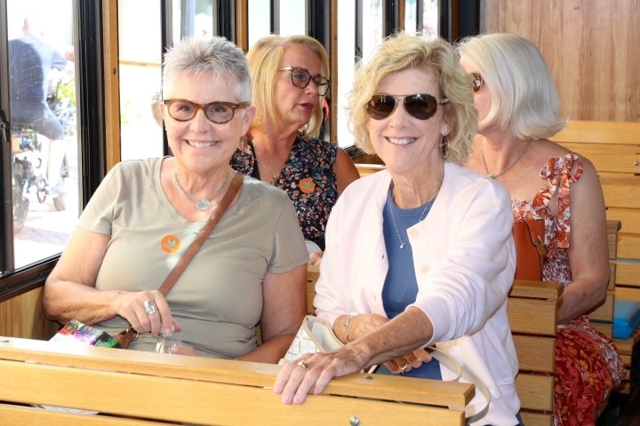 Five women sitting on a wooden bench inside a bus, smiling and wearing sunglasses.