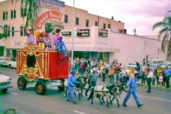 Parade with clowns on a red float pulled by miniature horses in a street with spectators.