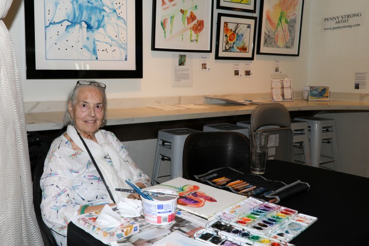 Elderly artist sitting at a table with watercolor paints and colorful artwork on the wall.