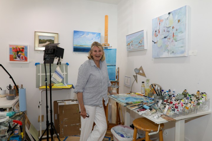 Person standing in an art studio with paintings and art supplies around.