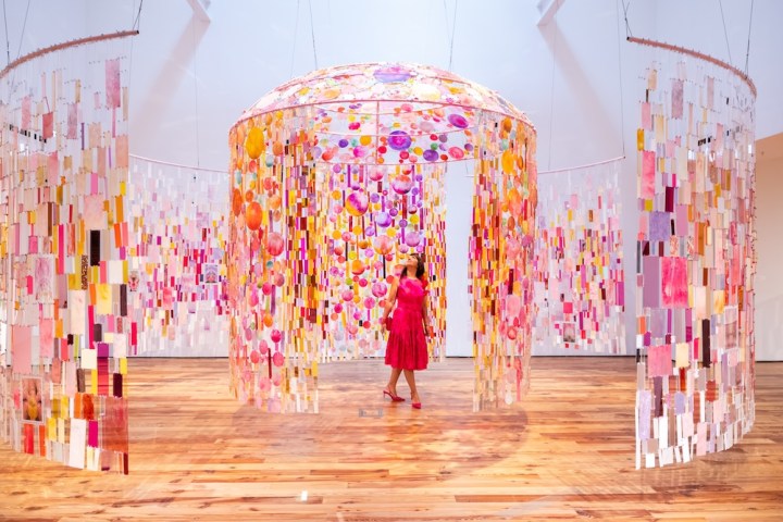 Woman in pink dress stands inside colorful hanging art installation.