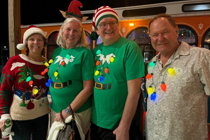Four people in Christmas attire, two wearing green shirts with elf designs, in front of a trolley at night.