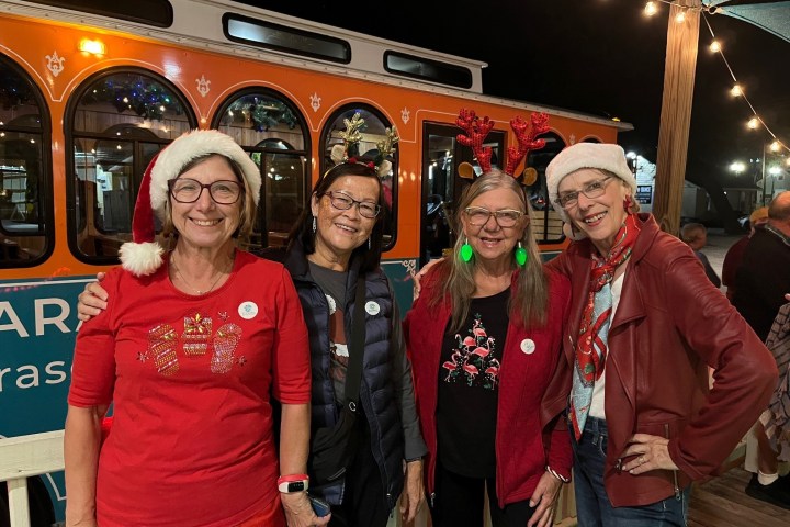 Four women in festive attire with Santa hats and antlers, smiling in front of an orange trolley at night.